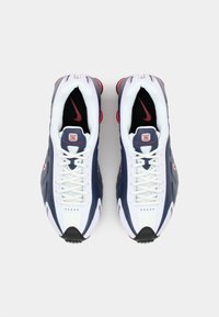 Nike Shox tenisice s bijelim gornjim dijelom, mornarsko plavim naglascima, crnim prednjim dijelom, crvenim logotipom i teksturiranim detaljima sa strane.