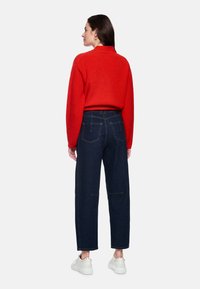 Roter, gerippter Pullover mit hohem Kragen, kombiniert mit hochtaillierten dunkelblauen Denim-Jeans und weißen Sneakers. Ansicht von hinten zeigt die Details des Outfits.