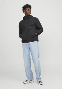 Black padded jacket with a zip, hood, and front pockets. Worn over a light purple shirt and paired with light blue jeans and sneakers.