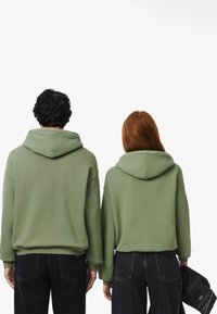 Felpe con cappuccio verdi realizzate in tessuto morbido, caratterizzate da una vestibilità comoda e polsini a coste. Indossate con jeans neri, viste da dietro.