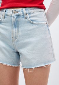 Ανοιχτό μπλε τζιν σορτς με ξεφτισμένο τελείωμα, φορεμένο από άτομο με κόκκινο μπλουζάκι και μακρύ λευκό μανίκι, εμφανιζόμενο από τη μέση έως το μέσο μηρού.