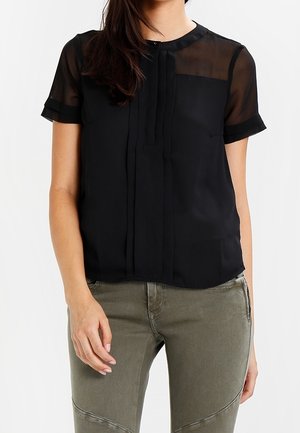 Kvinde iført en sort gennemsigtig kortærmet bluse med lodrette plisseringer, kombineret med olivengrønne tætsiddende bukser.