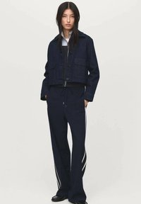 Marineblauwe denim cropped jack met twee voorzakken, gecombineerd met marineblauwe joggingbroeken met witte zijstripes en een elastische tailleband.