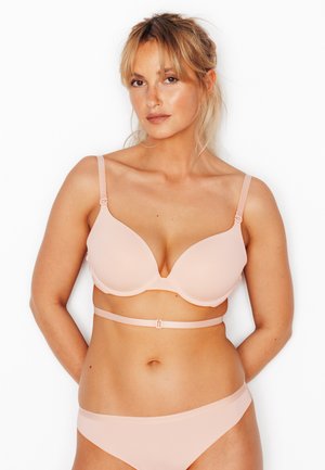 Femme aux cheveux blonds portant un soutien-gorge rembourré rose clair et une culotte assortie, debout avec les bras derrière le dos sur fond blanc.