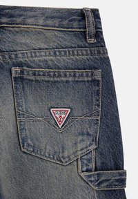 Kieszonka z denimu w niebieskim odcieniu i widocznym przeszyciem. Zawiera trójkątną łatkę z logo "GUESS" w kolorze czerwonym i białym na tylnej kieszeni.