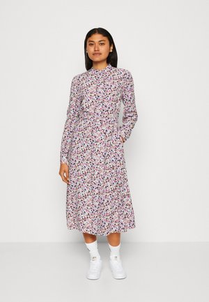 Florales Midi-Kleid mit langen Ärmeln, weißem Hintergrund, buntem Blütenmuster in kleinen Größen und taillierter Passform. Das Modell trägt weiße Sneaker.