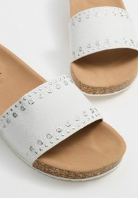 Chanclas blancas con una parte superior texturizada, decoradas con tachuelas plateadas y una suela de corcho. Diseño plano con una plantilla cómoda.