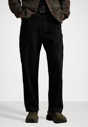Black corduroy pants with a relaxed fit and side pockets, paired with olive green outdoor shoes. Texture is soft, and the belt is black.