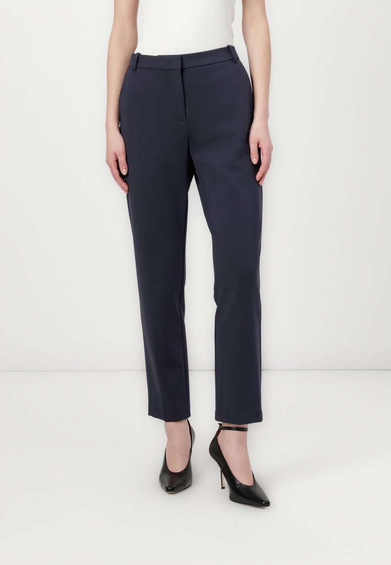 Mornarsko modre prilagojene hlače z ožjim fitom, gladkim materialom, zankami za pas in stranskimi žepi. Uporabljene s črnimi čevlji na visoki peti.