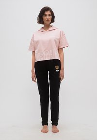 Woman standing barefoot wearing a pink cropped hoodie and black sweatpants with a teddy bear logo.