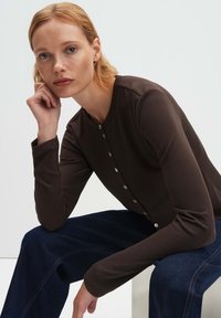 Frau mit hellrotem Haar, die ein braunes, mit Knöpfen versehenes Langarmoberteil und dunkelblaue Hosen trägt, sitzt und stützt ihren Kopf auf ihre Hand.