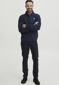 Marinefarbener Fleece-Pullover mit Reißverschlusskragen, ausgestattet mit einem Logo-Patch auf der linken Brust und seitlichen Reißverschlusstaschen, kombiniert mit dunkler Hose und schwarzen Stiefeln.