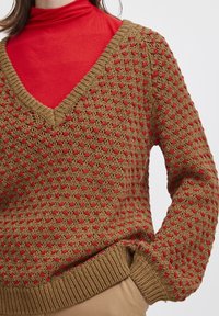 Pull en tricot avec un col en V marron, présentant des accents rouges et un motif texturé. Ourlet et poignets côtelés. Superposé sur un col roulé rouge.
