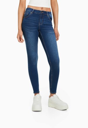 Frau trägt dunkelblaue Skinny-Jeans, weißes Croptop und weiße Plateausneaker, steht vor weißem Hintergrund.