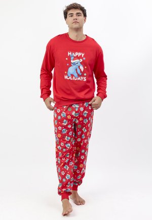 Joven con pijama roja temática navideña que presenta un personaje azul con un gorro de Santa y el texto "Felices Fiestas", de pie descalzo.