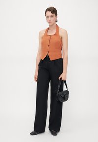 Orange halter neck vest with button front, paired with black wide-leg trousers. Model holds a small black crossbody bag and wears studded flats.