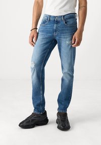 Vaqueros de mezclilla azul con un corte relajado, que presentan detalles de desgaste en las rodillas y un diseño clásico de cinco bolsillos, combinados con zapatillas deportivas negras.