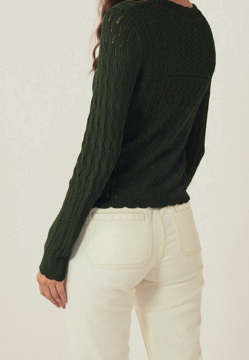 Femme portant un pull à manches longues texturé vert foncé et un jean taille haute blanc cassé, vue de dos et de la taille vers le haut.