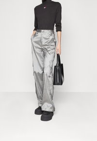 Person som bär glänsande silver cargo-byxor, svart långärmad tröja med liten logotyp, svarta platåskor och håller en svart handväska.