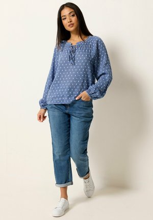 Joven mujer de pie con una blusa azul de manga larga con estampado, vaqueros azules remangados y zapatillas blancas sobre un fondo claro liso.