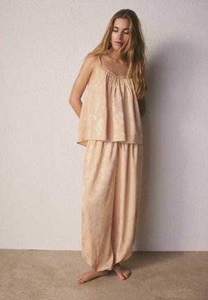 Jeune femme aux longs cheveux portant un haut beige ample sans manches et un pantalon large assorti, debout pieds nus contre un mur blanc texturé.