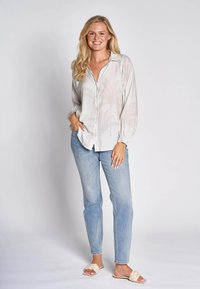 Hellgraue Bluse mit Palmenblattmuster, durchgehender Knopfleiste und legeren Ärmeln, kombiniert mit hellblauen Jeans und beigen Slide-Sandalen.
