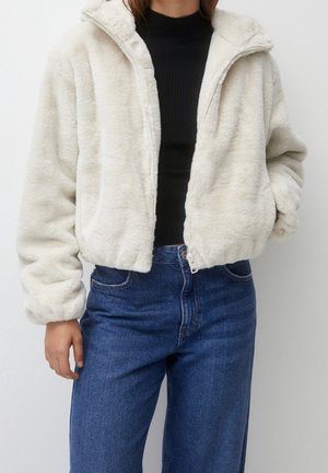 Femme portant une veste blanche duveteuse zippée par-dessus un haut côtelé noir avec un jean taille haute bleu sur un fond uni.