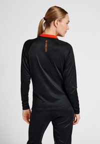 Maglietta sportiva a maniche lunghe nera con colletto rosso a contrasto e dettaglio del logo. Tessuto liscio, design aderente, stile minimalista.