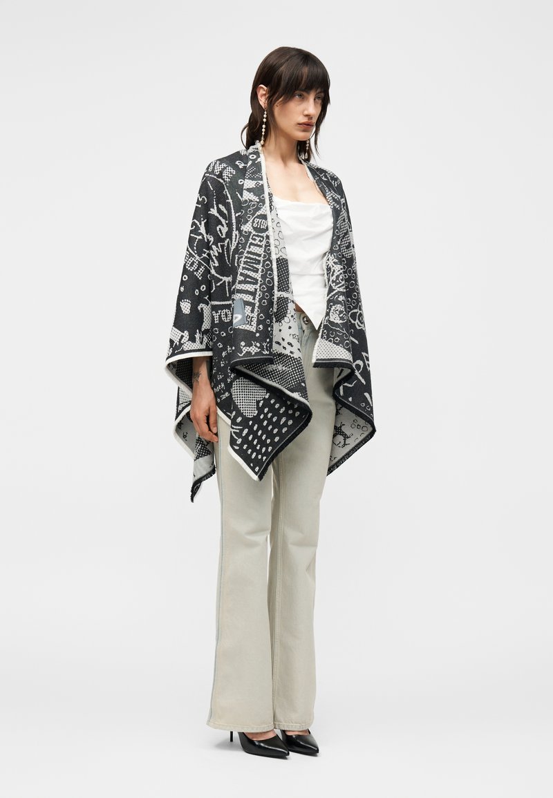 Femme portant un châle à motif noir et blanc sur un haut sans manches blanc, pantalon large beige clair et talons noirs pointus, debout.