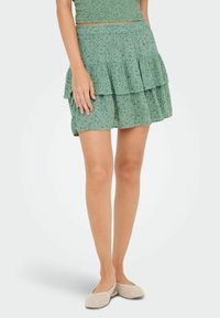 Mujer con una falda verde con flores en capas y zapatos tipo slip-on de ratán en color beige, de pie con una mano apoyada en su falda.