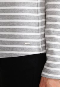 Maglia a maniche lunghe a strisce grigie e bianche con una texture liscia, dotata di un piccolo etichetta in metallo argentato vicino all'orlo. Pantaloni scuri parzialmente visibili.
