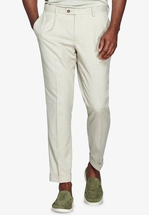 Hombre con pantalones beige a medida con puños, manos en los bolsillos y zapatos tipo mocasín de ante verde oliva, caminando hacia adelante.