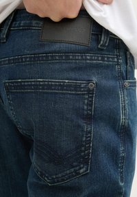 Los vaqueros de mezclilla azul oscuro presentan una tela texturizada, un parche de cuero con el logo en la cintura, y acentos de bolsillo trasero con doble costura.