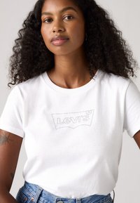 Balta medvilnė marškinėliai su siuvinėtu Levi's logotipu sidabrinės spalvos. Klasikinis apvalus iškirpimas ir trumpomis rankovėmis, derinami su aukštu liemeniu džinsais.