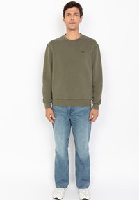 Oliwkowy bawełniany sweatshirt z okrągłym dekoltem i ściągaczami, w połączeniu z jasnoniebieskimi dżinsami prostymi i białymi sneakersami.