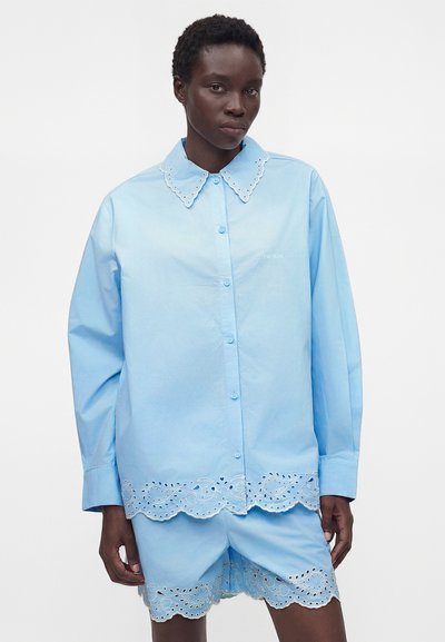 Μοντέλο που φοράει ανοιχτό μπλε πουκάμισο με κουμπιά και σορτς με κυματιστή δαντέλα με οπές στο γιακά, το στρίφωμα και το άνοιγμα των ποδιών.