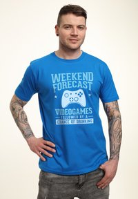 Modré bavlněné tričko s nápisem "PREDIKCE NA VÍKEND: VIDEOHRY, NÁSLEDOVANÉ ŠANCÍ NA PITÍ." Krátké rukávy a kulatý výstřih.