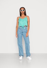 Top sem mangas verde menta com botões e bainha amarrada, combinado com jeans azuis de cintura alta e ténis brancos. Fundo liso.