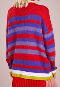 Kolorowy sweter z wyraźnymi czerwonymi, fioletowymi i szarymi poziomymi pasami. Dzianina o fakturze, luźny krój i ściągacz na dole w kolorach zielonym i białym.