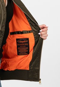 Giubbotto bomber khaki con fodera arancione; presenta una cerniera, polsini a coste e un'etichetta nera con dettagli sul marchio e le istruzioni per la cura.
