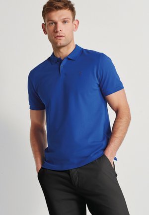 Camisa polo azul de tela suave, con cuello clásico, mangas cortas y un pequeño logo bordado en el pecho. Pantalones negros.