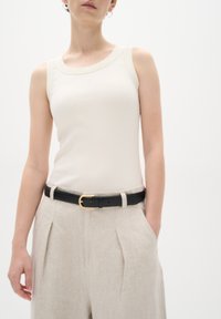 Ljusbeige ribbad tanktop, svart bälte med guldfärgad spänne, och lösa ljust krämfärgade linbyxor med veck, på en vit bakgrund.