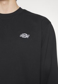 Personne portant une chemise noire à manches longues avec un petit logo blanc brodé "Dickies" sur la poitrine gauche sur fond uni.