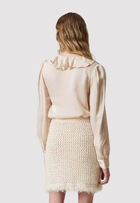 Blusa beige chiaro con colletto arricciato e maniche lunghe abbinata a una gonna crema strutturata con macchie colorate e orlo frangiato.