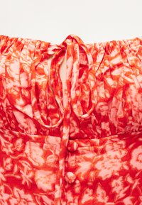 Κόκκινο και λευκό φόρεμα με λουλουδάτο μοτίβο, με σούρες στη λαιμόκοψη, υφασμάτινους δεσμούς και κουμπιά καλυμμένα με ύφασμα κατά μήκος του μπροστινού μέρους.