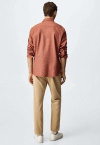 Raskorven paita, jossa on käännetyt hihat ja suora leikkaus, yhdistettynä vaaleisiin housuihin ja valkoisiin lenkkareihin vaalealla taustalla.