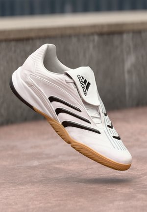 White athletic shoe with black accents and textured grooves, featuring a round toe and a gum sole. Logo on the tongue.