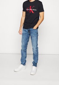 Svart bomullströja med röd och vit "Calvin Klein Jeans"-logotyp, kombinerad med ljusblå denimjeans och vita sneakers.