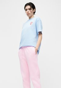 Νεαρό άτομο με κοντά σκουρόχρωμα μαλλιά, που φοράει ένα ανοιχτό μπλε μπλουζάκι και ανοιχτό ροζ φόρμα, στέκεται με τα χέρια στις τσέπες.