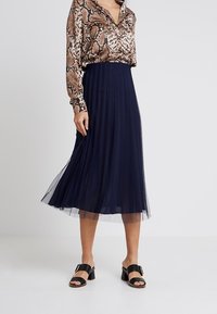 Gonna midi a pieghe blu navy realizzata in tulle leggero, abbinata a una blusa marrone con stampa serpente e sandali neri con tacco.